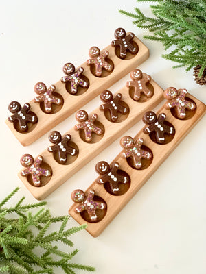 Three wooden five frame sensory trays with decorated gingerbread man-shaped loose parts on white surface with pine branches