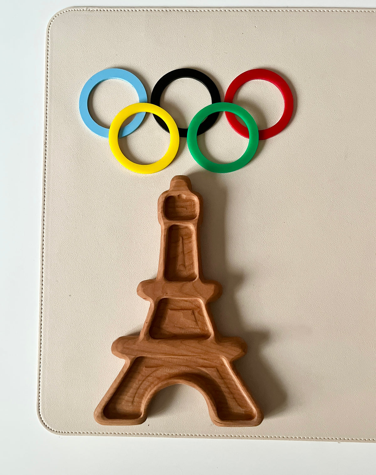 Wooden tray carved in the shape of the Eiffel Tower below five colorful interlocking rings on a beige surface