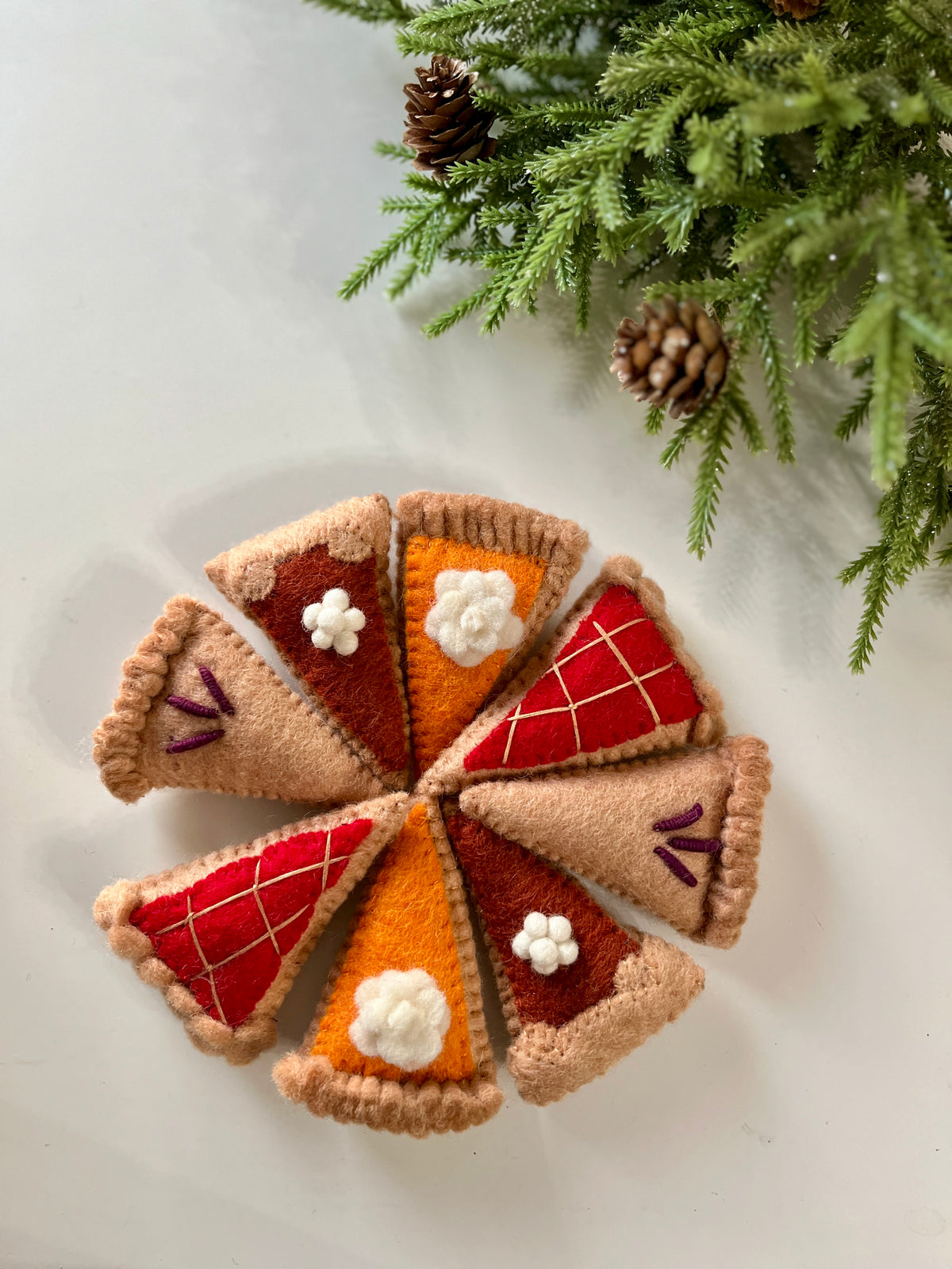 Eight felt pie slices with stitched crusts and colorful toppings arranged in a circle near pine branches and cones
