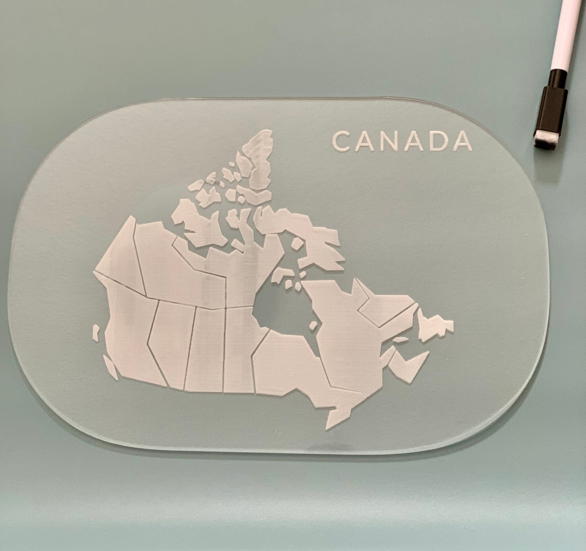Clear map of Canada with provincial borders engraved on an oval translucent light green surface next to a stylus