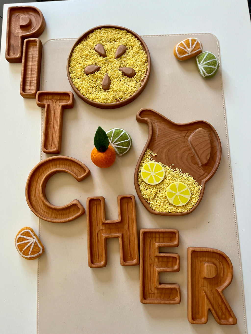 Wooden letter trays spelling "Pitcher" with a wooden pitcher and plate holding yellow rice and fruit decorations on a beige mat