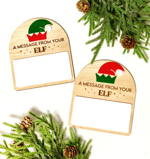 Two wooden note holders with elf hat designs and the text "A message from your ELF" surrounded by pine branches and cones