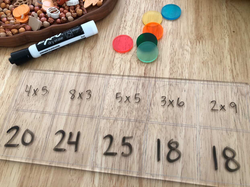 Transparent board with multiplication problems and answers, colorful plastic counters, dry erase marker, and sensory play materials nearby