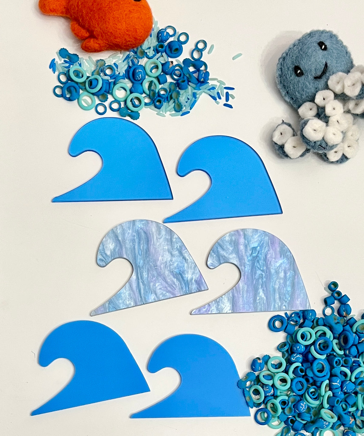 Six wave-shaped acrylic pieces in solid blue and marbled blue tones surrounded by blue confetti and plush toys