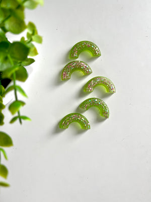 Five translucent lime green resin half-circle pieces embedded with pink and silver glitter on a white surface near green leaves