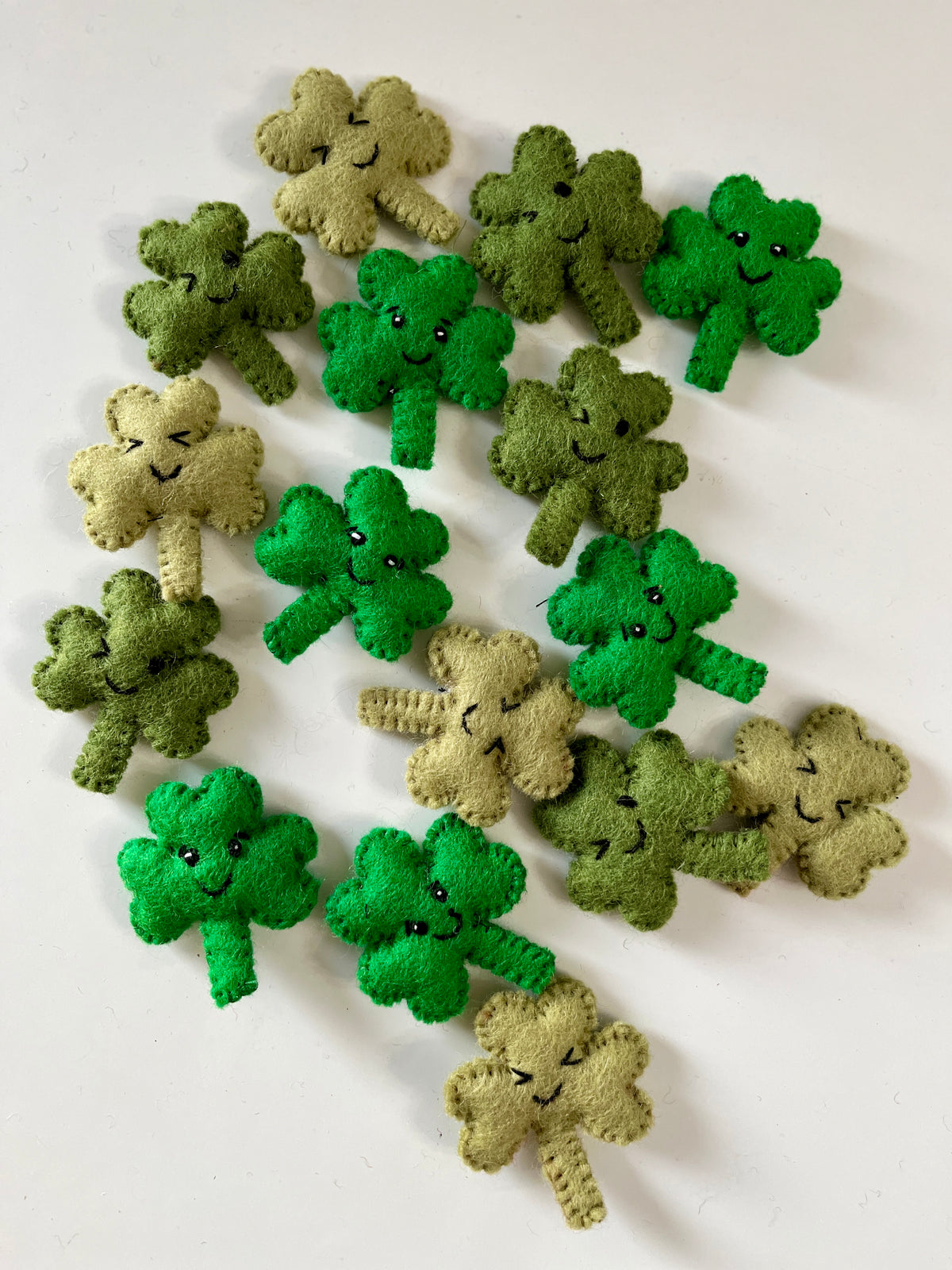 Assorted small felt four-leaf clover shapes in shades of green and beige with stitched smiling faces on a light background