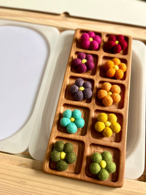 Wooden tray with ten compartments holding colorful felt flowers in shades of pink, orange, purple, blue, yellow, and green on a wooden surface