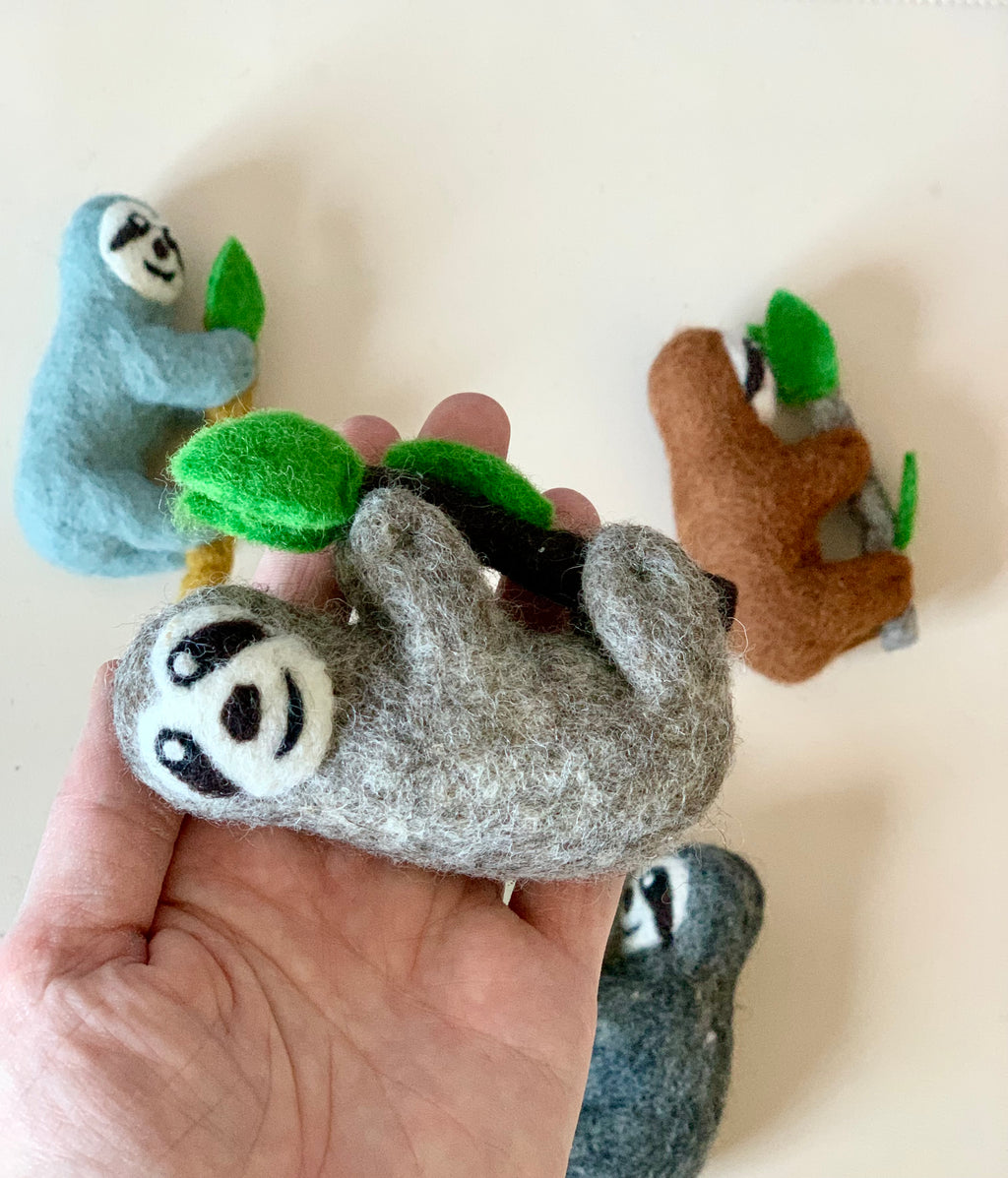 Hand holding a gray felt sloth figurine clinging to a branch with green leaves with three other felt sloths in different colors nearby
