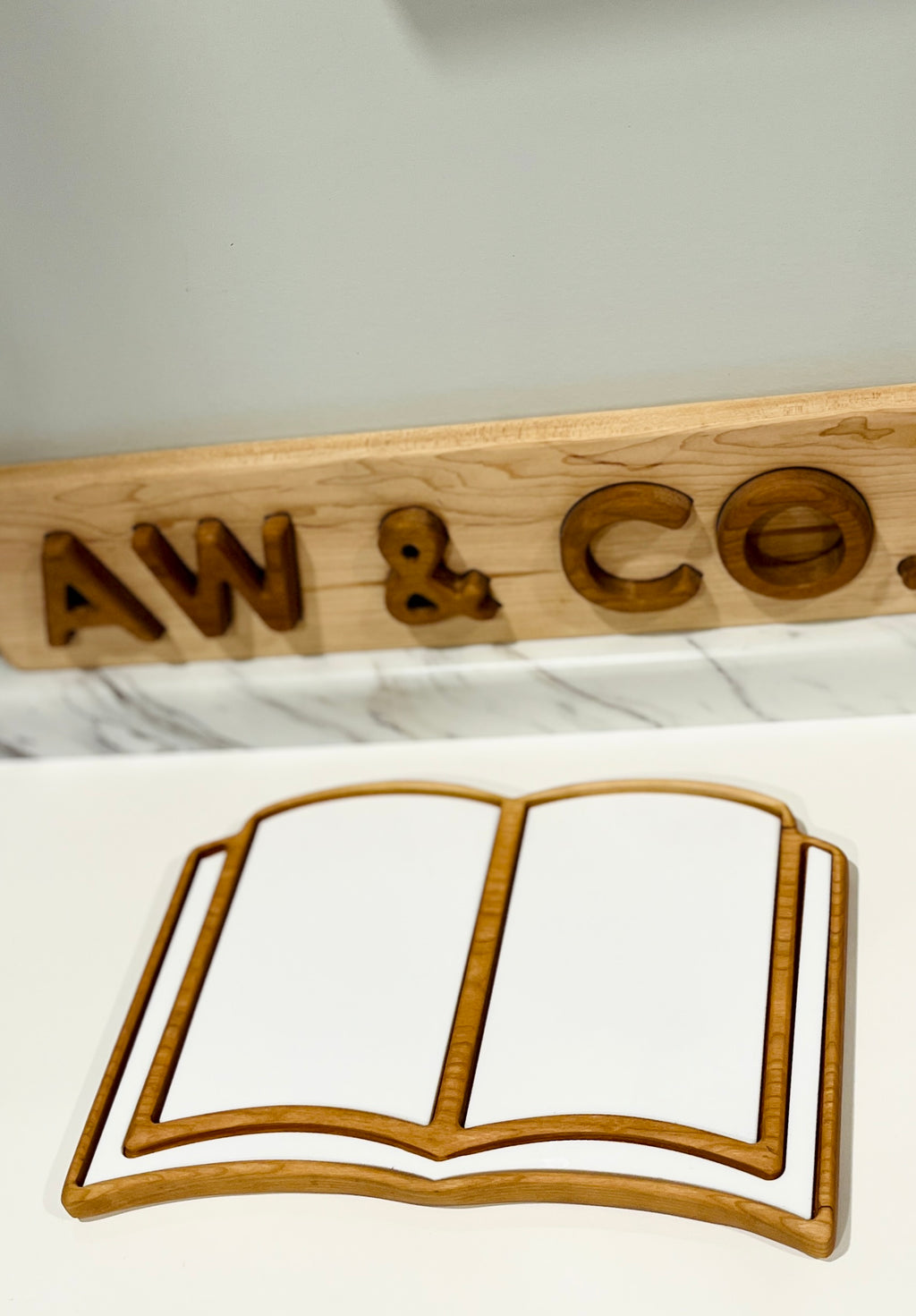 Thin wooden tray shaped like an open book placed on a white surface with a carved wooden sign behind it