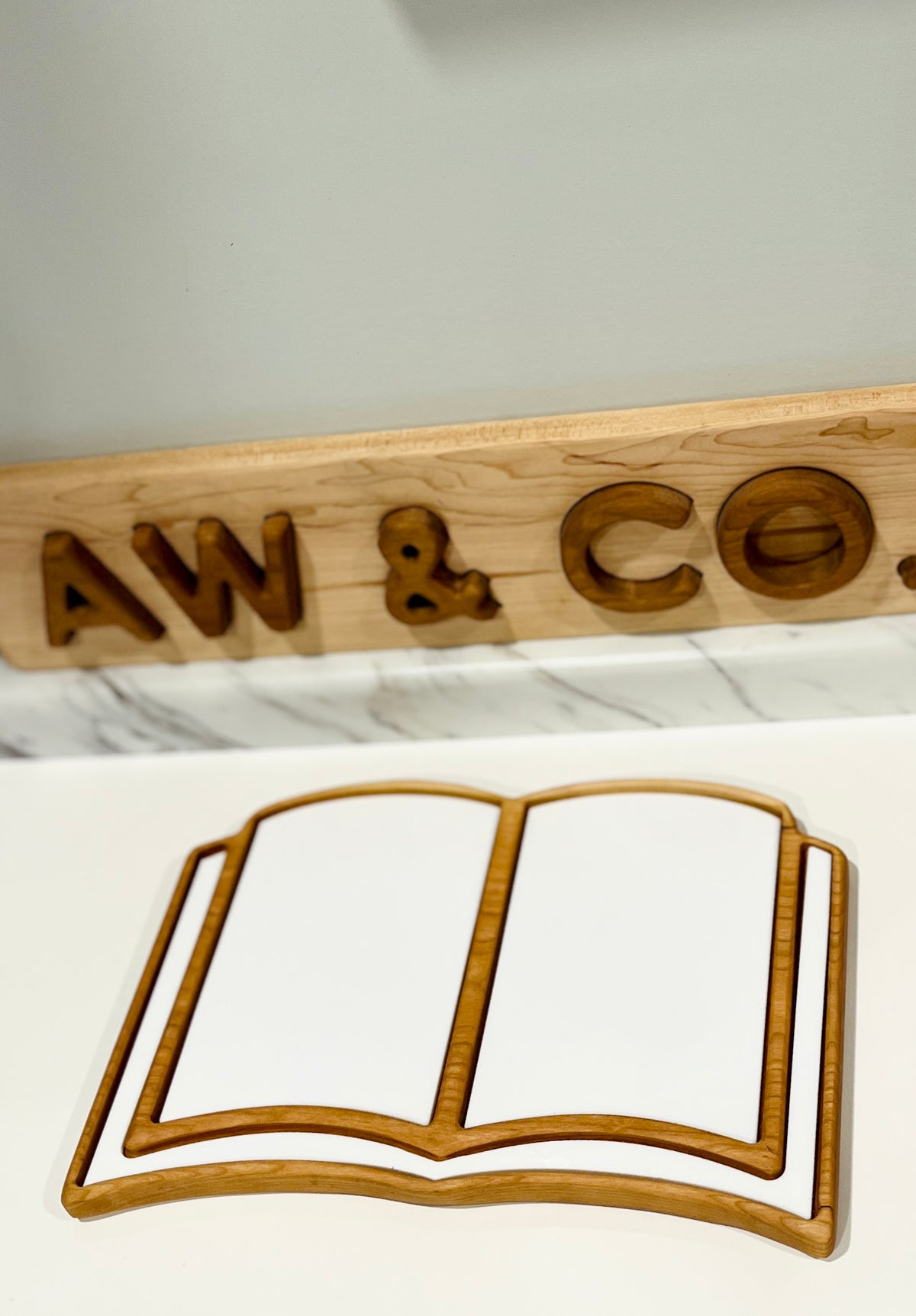 Thin wooden tray shaped like an open book placed on a white surface with a carved wooden sign behind it