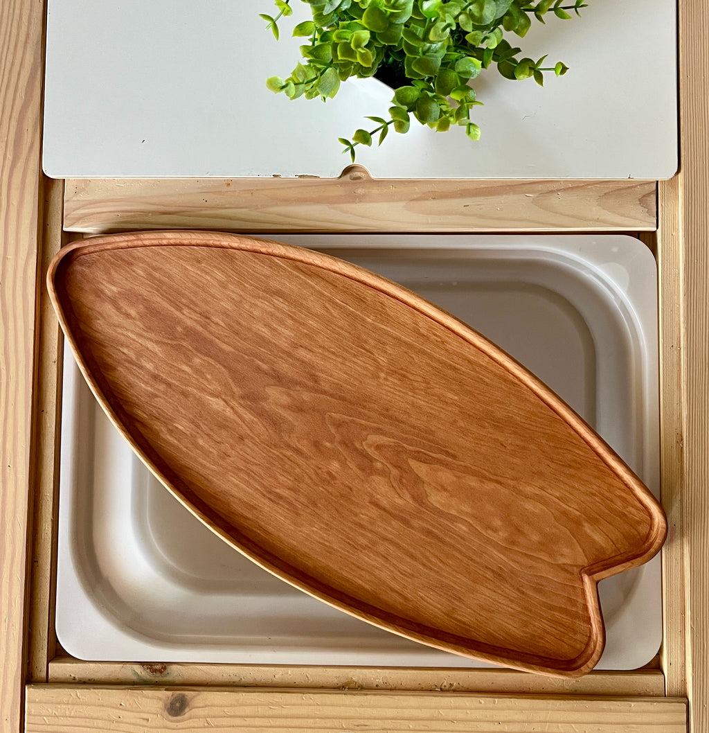 Smooth wooden surfboard-shaped tray resting inside a recessed wooden frame with a green leafy plant above