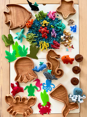 Wooden ocean creature-shaped trays with colorful sensory pasta, shells, felt sea animals, and silicone seaweed on a light surface