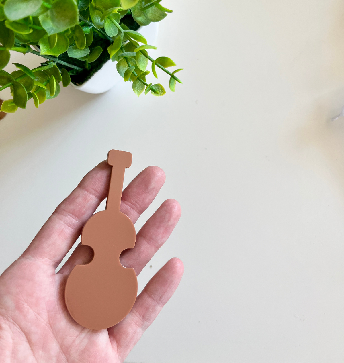 Hand holding a smooth matte light brown violin-shaped cutout with green leaves of a potted plant on a white surface