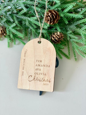 Light wood arch ornament engraved with family names, "Christmas 2023," and hanging from twine against pine branches and cones