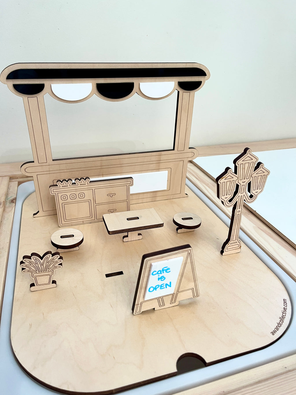 Laser-cut wooden miniature café scene with table, stools, streetlamp, potted plant, and signboard on a light wood base