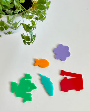 Five colorful acrylic shapes including green frog, orange fish, blue carrot, purple flower, and red crane on white surface with plant leaves nearby