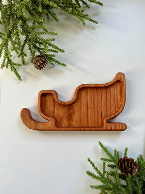 Wooden tray carved in the shape of a sleigh surrounded by pine branches and pine cones on white surface