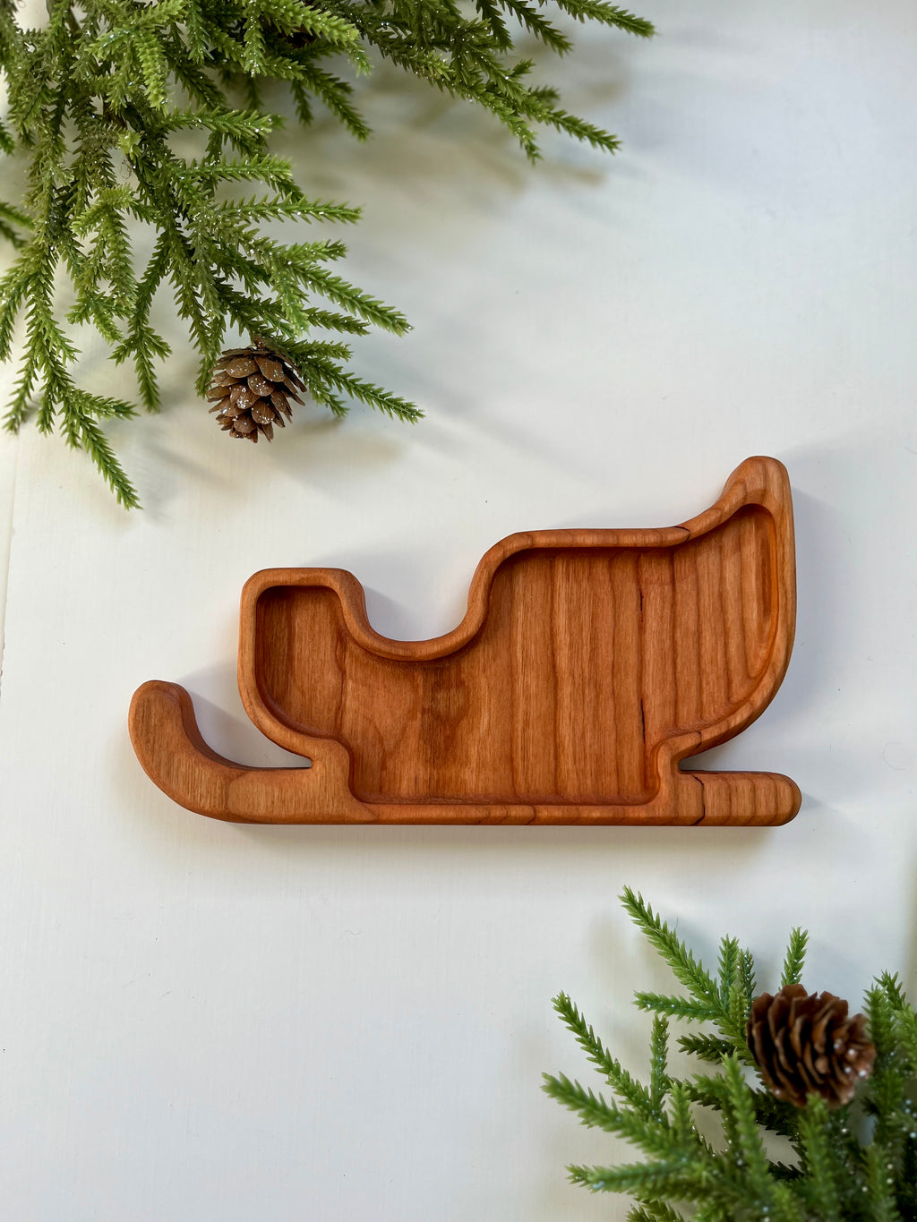 Wooden tray carved in the shape of a sleigh surrounded by pine branches and pine cones on white surface