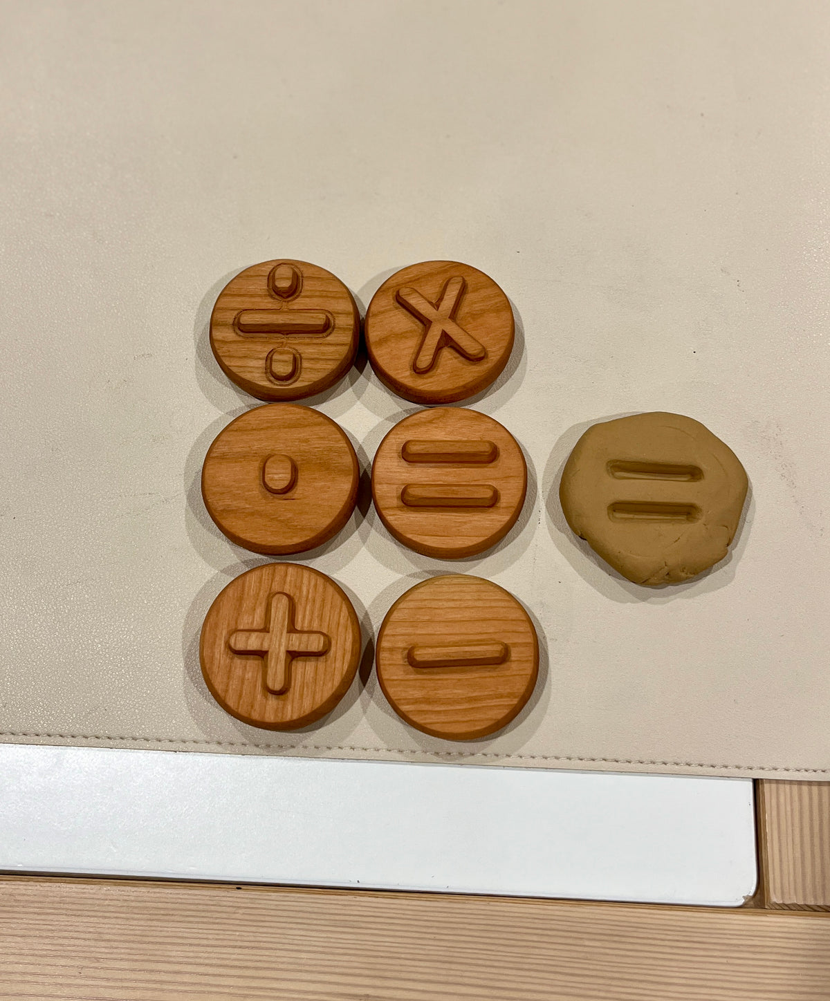 Six round wooden tokens with engraved math symbols and a clay piece imprinted with an equal sign on a pale surface
