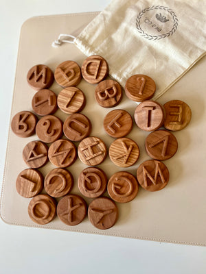 Round wooden tokens engraved with uppercase letters scattered beside a beige drawstring pouch on a light surface