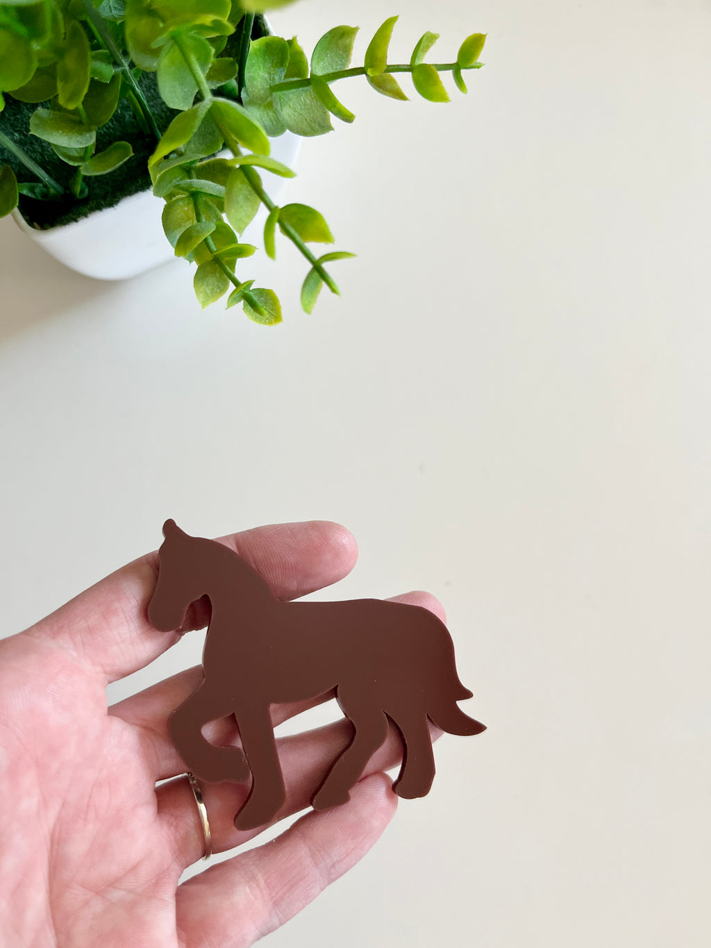 Hand holding a smooth brown horse-shaped cutout with a small green potted plant in the background
