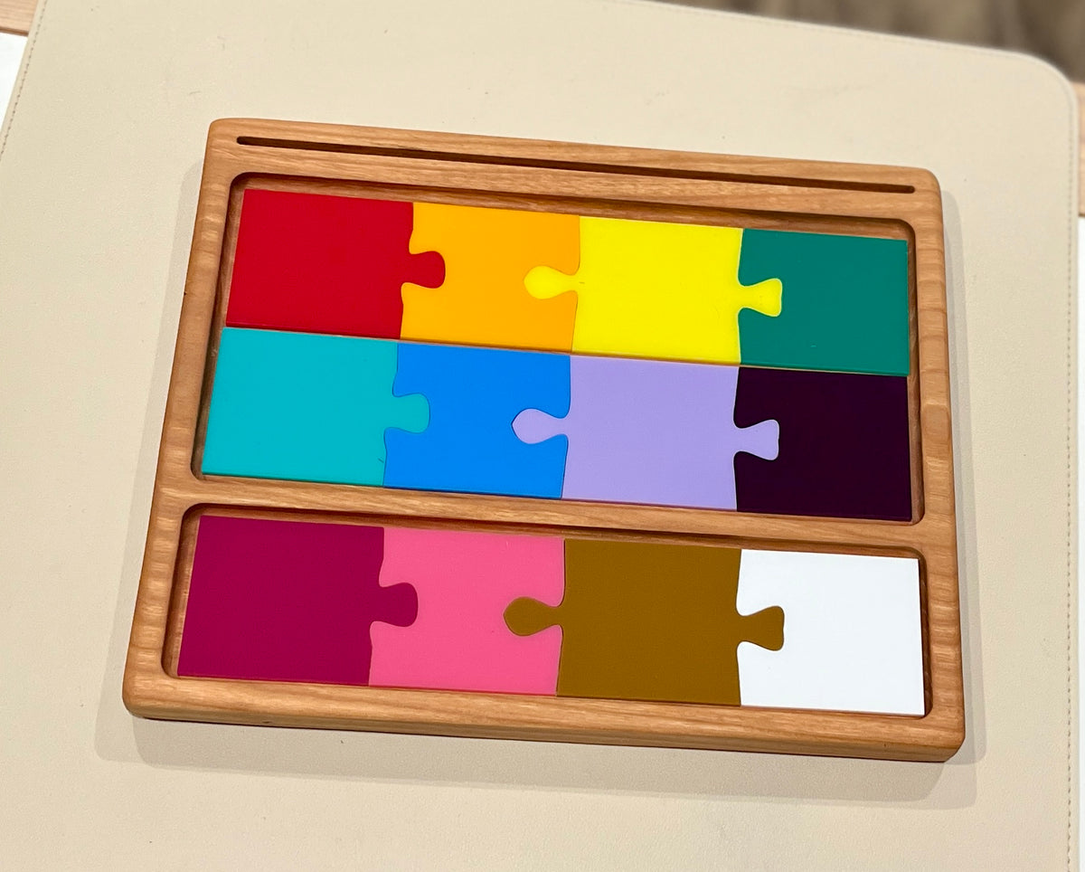 Colorful rectangular wooden puzzle pieces in a light wood tray arranged in three rows on a light background