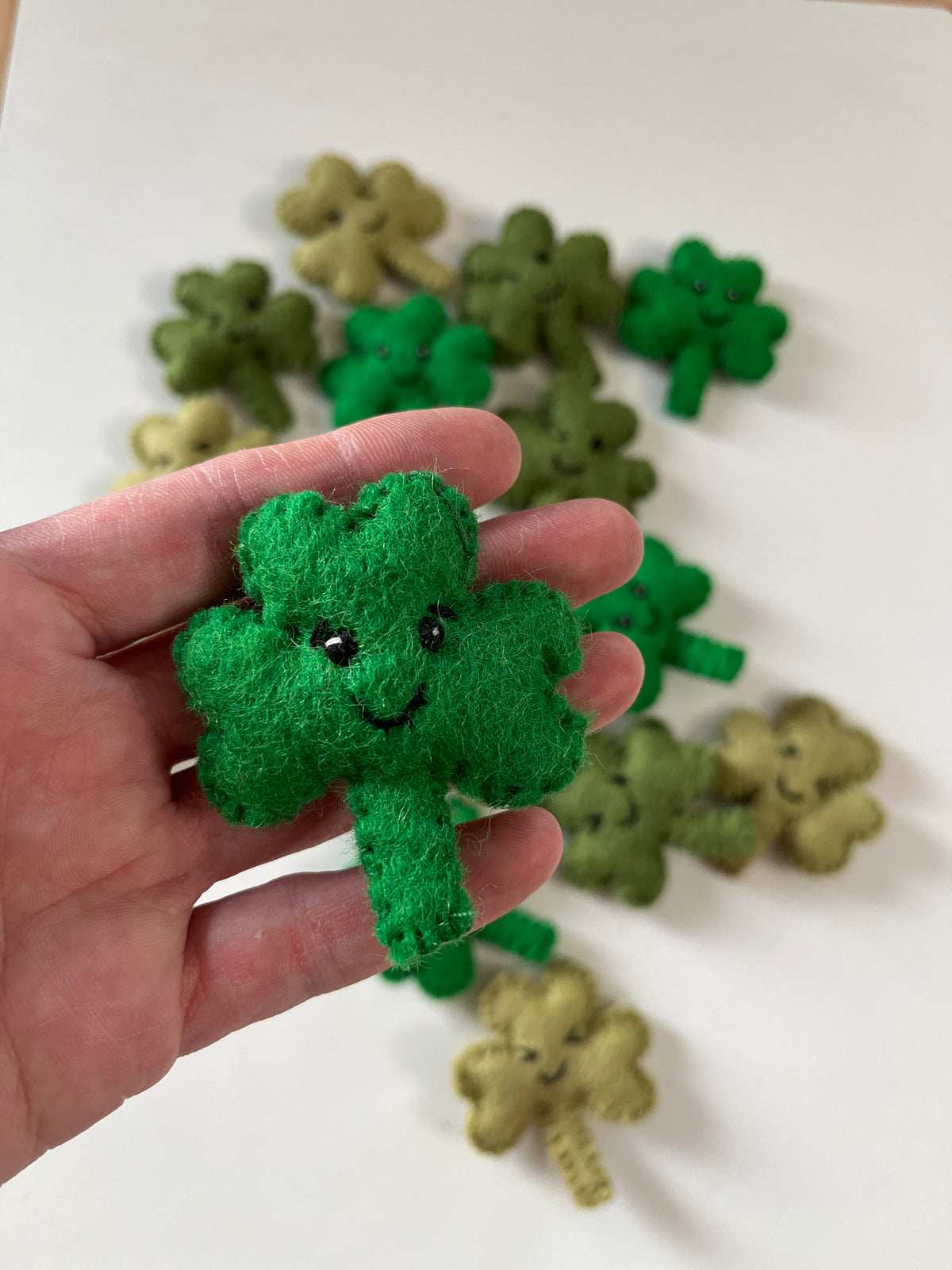 Hand holding a small green felt shamrock with a smiling face, multiple similar shamrocks in varying greens in the background