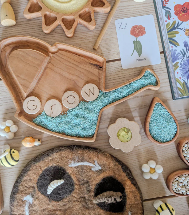 Wooden watering can sensory tray filled with blue rice, wooden letter discs spelling grow, and nature-themed toys on wooden table