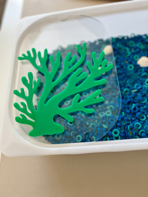 Green coral-shaped plastic decoration on clear lid of white tray filled with small blue circular beads and white shells