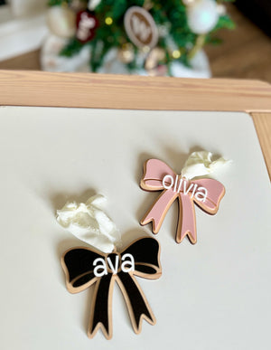 Two wooden bow ornaments on white surface, one black with name "ava" and one pink with name "olivia" attached with white ribbons