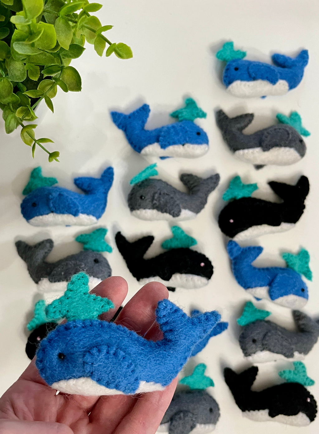 Hand holding a small blue and white felt whale with turquoise water spout, surrounded by similar whales in blue, gray, and black on white surface