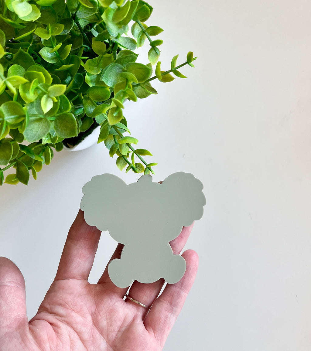Hand holding a smooth pale green koala-shaped cutout beside a vibrant green potted plant on a light surface