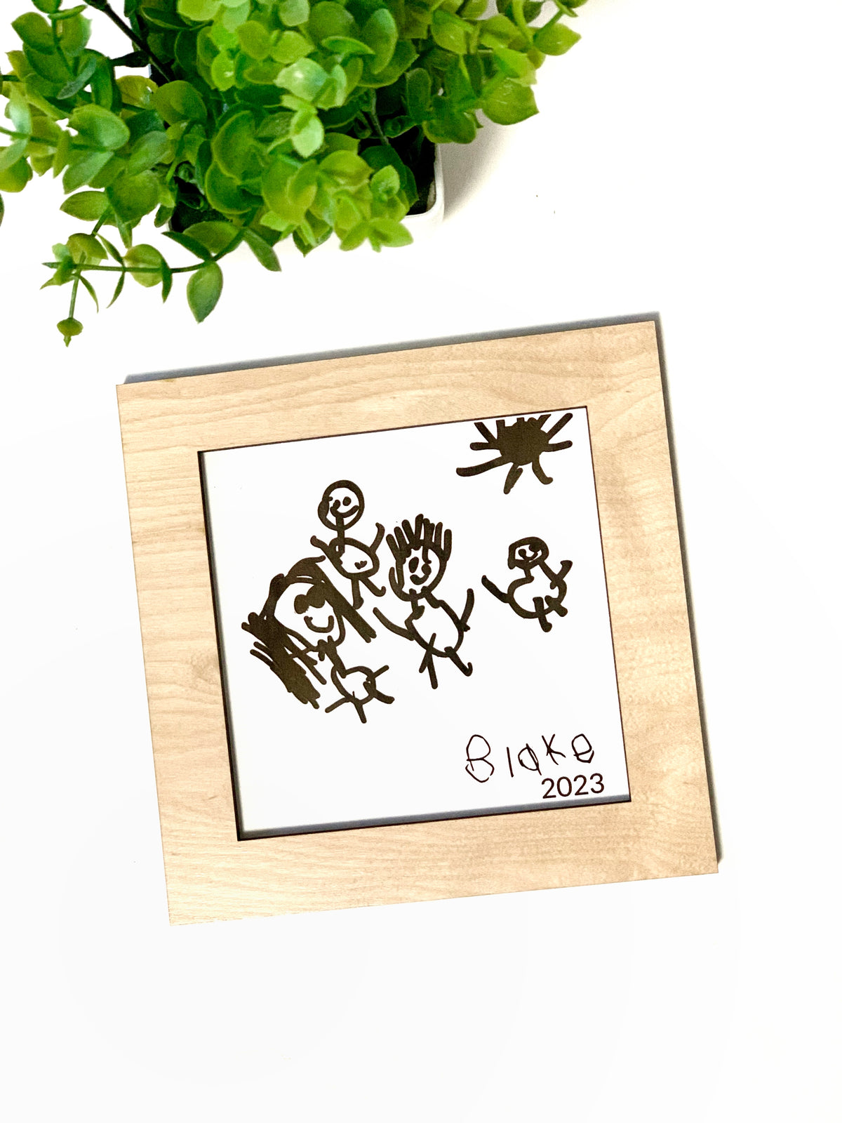 Child’s drawing traced in black ink on white paper, framed in natural wood square with green plant in background