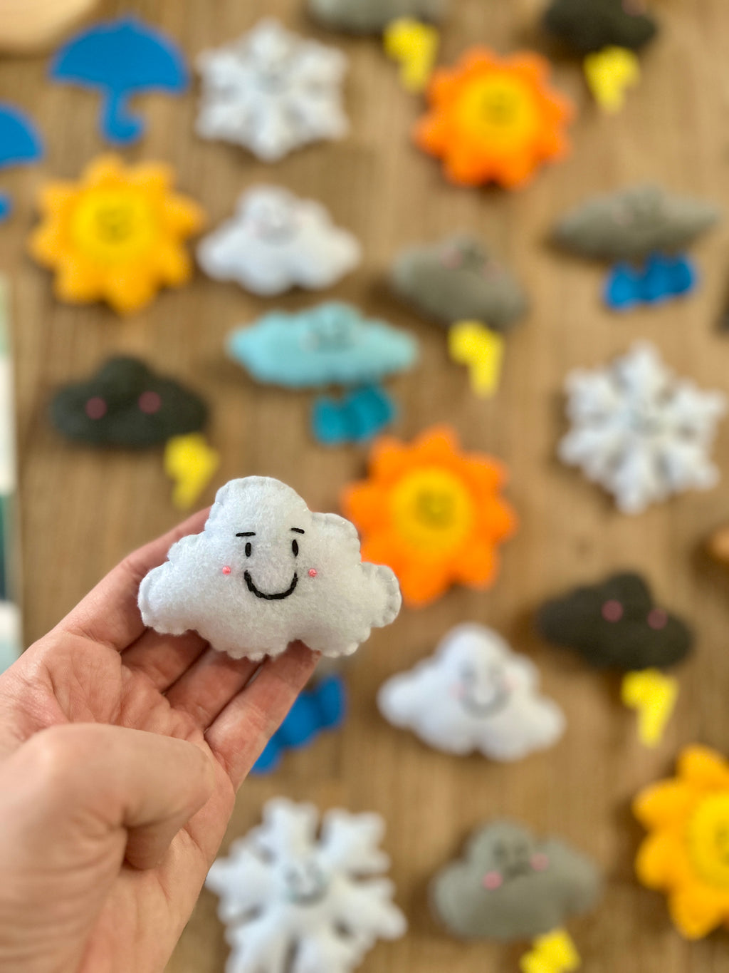 Hand holding a smiling white felt cloud with embroidered face, with various colorful Felt Weather Shapes, Made in Canada, handcrafted, homeschool, education resources scattered on a wooden surface.