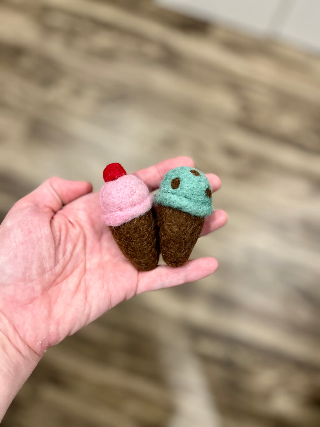 Hand holding two small felt ice cream cones, one pink with a red cherry and one mint green with brown spots, wooden floor background