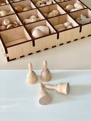Four smooth unfinished wooden bell shapes on a white surface with a wooden compartment tray holding various wooden pieces in the background