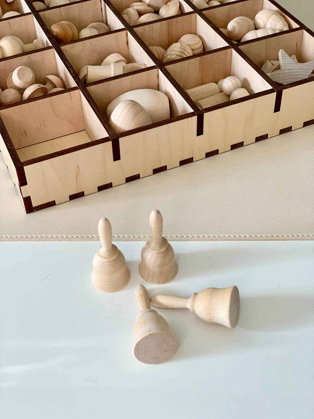 Four smooth unfinished wooden bell shapes on a white surface with a wooden compartment tray holding various wooden pieces in the background