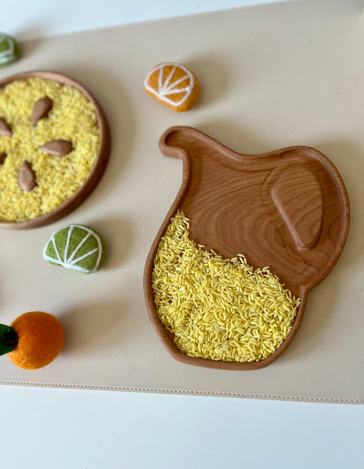 Wooden pitcher-shaped tray partially filled with yellow rice grains surrounded by felt fruit slices on a beige mat
