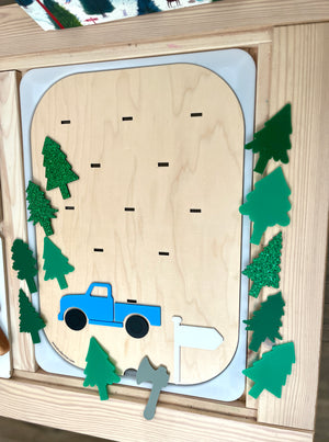 Wooden activity board with blue truck, white flag, gray axe, and various green tree shapes with some glitter details