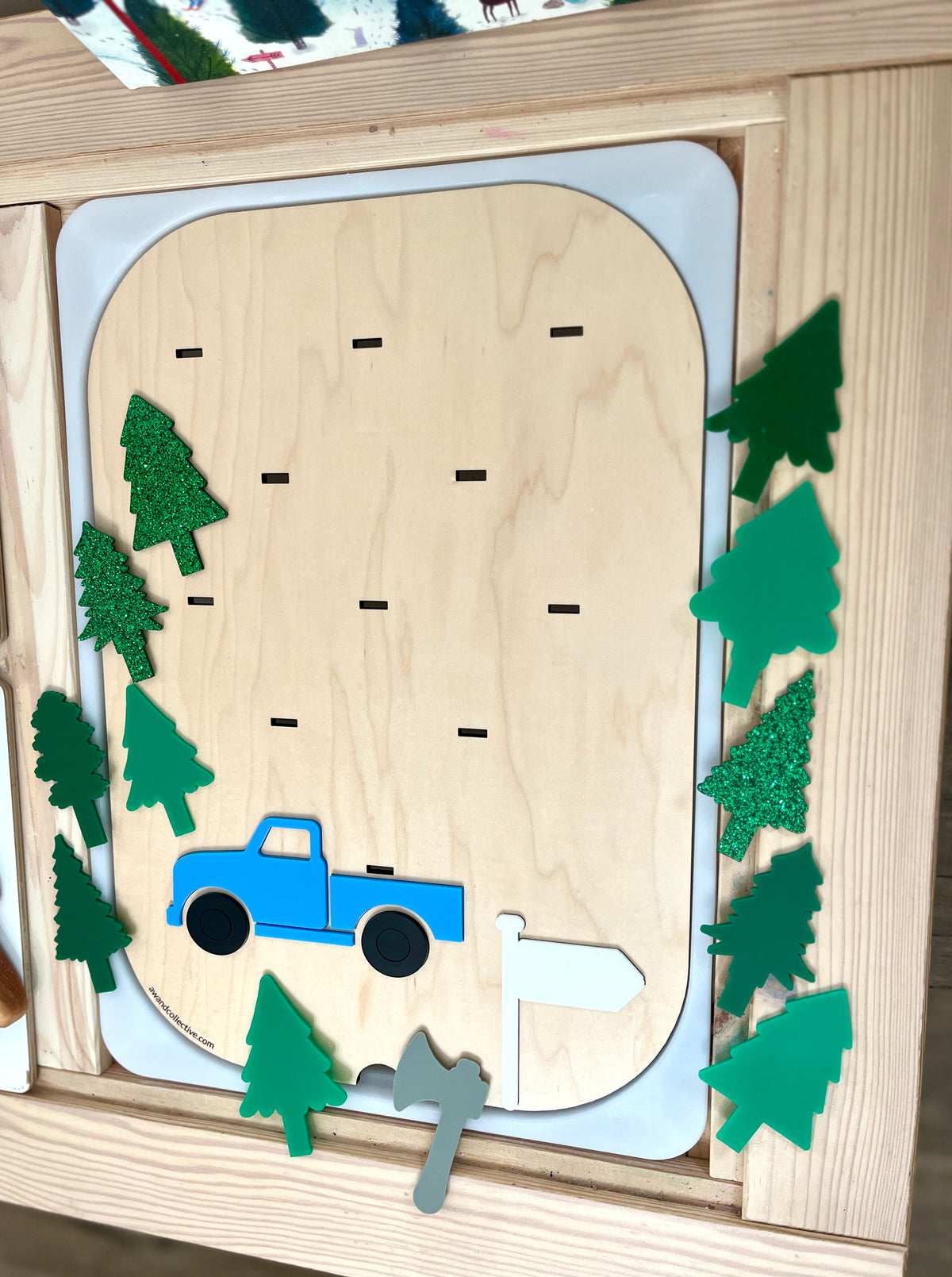 Wooden activity board with blue truck, white flag, gray axe, and various green tree shapes with some glitter details