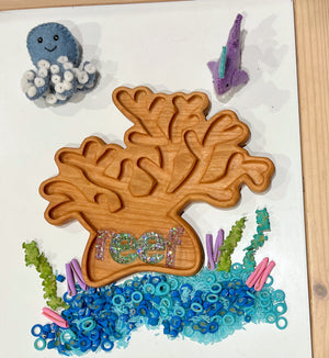 Wooden coral-shaped tray filled with blue and green sea-themed beads and surrounded by felt sea creatures on white surface
