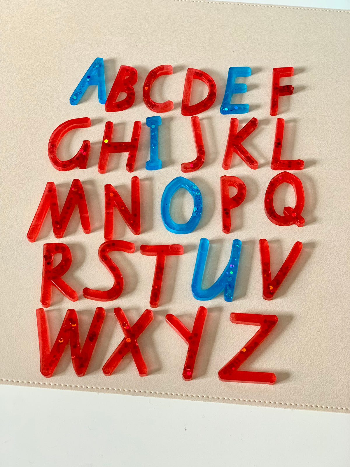 Translucent resin alphabet letters in red and blue colors arranged in five staggered rows on a beige surface