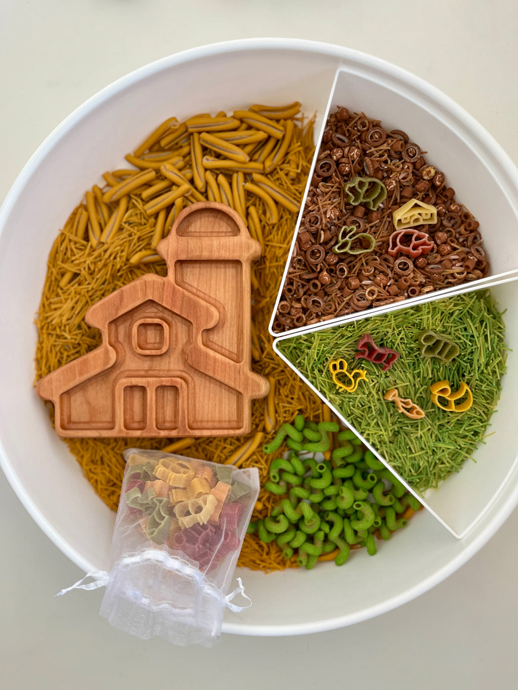 Round white tray with sections of yellow, green, and brown pasta, wooden barn scoop, and small colorful pasta pieces in mesh bag