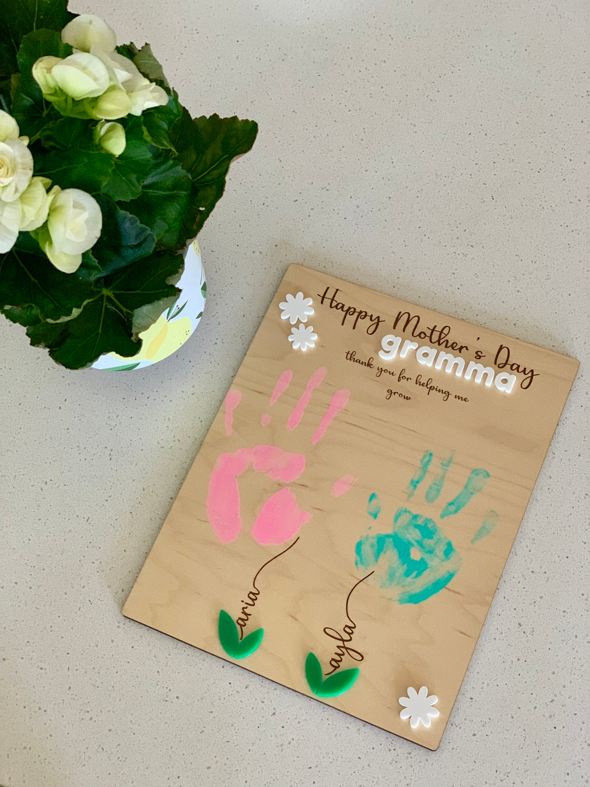 Wooden plaque with pink and blue handprints, green leaves, white flowers, and Mother's Day message for grandma on a beige surface
