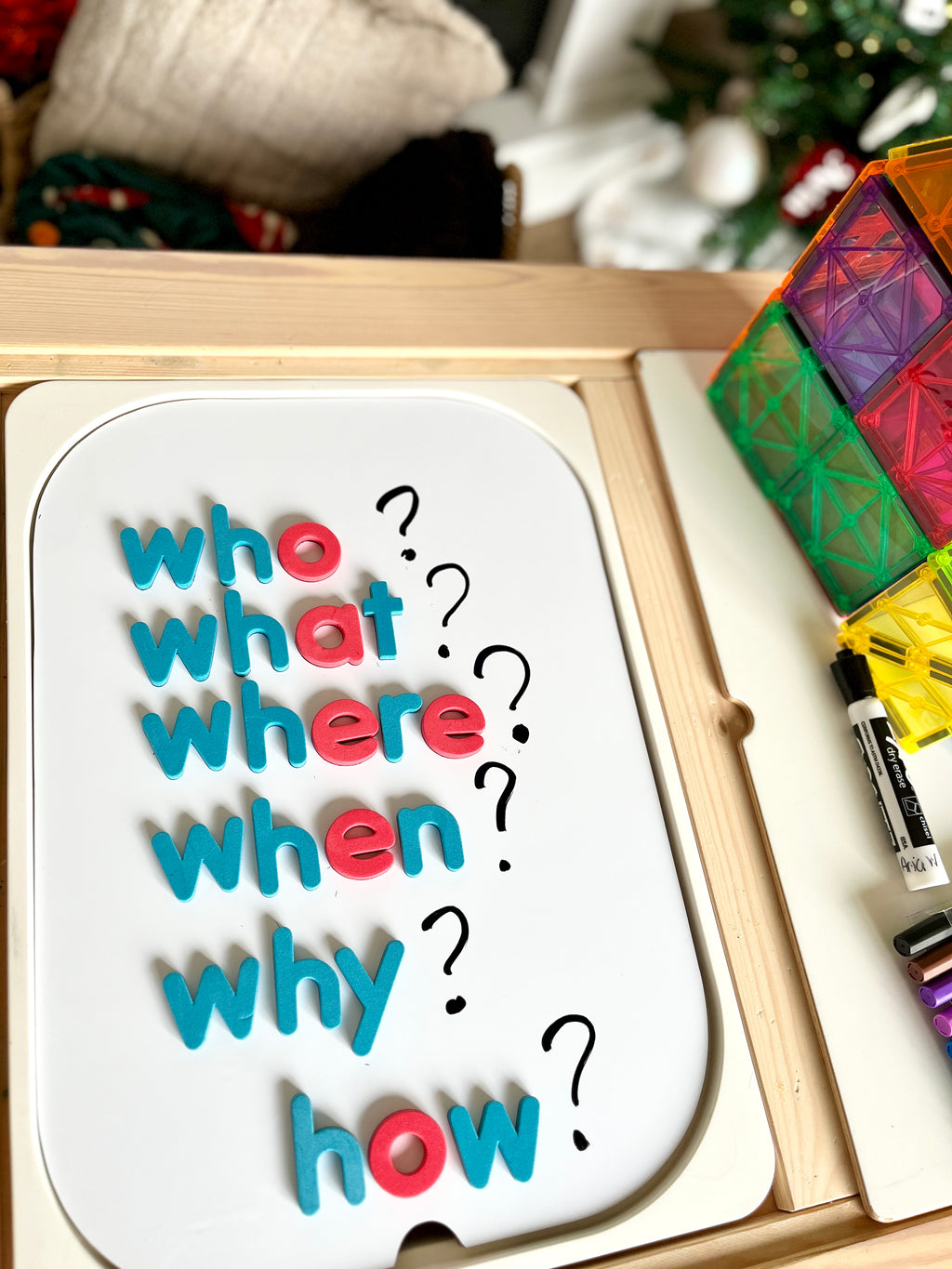 White magnetic board with blue and red letters spelling question words accompanied by black question marks and colorful tiles nearby
