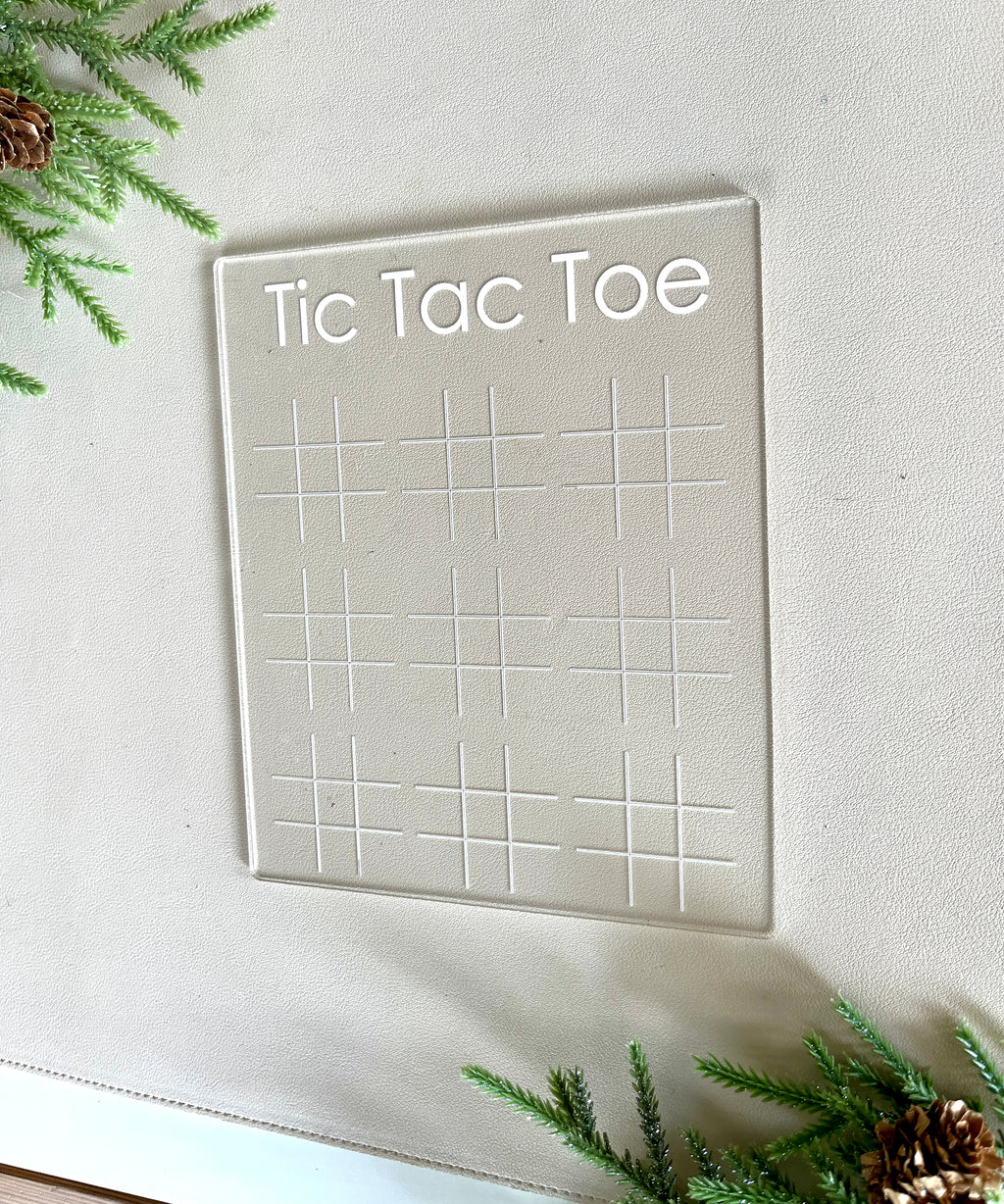 Transparent acrylic Tic Tac Toe game board with engraved grids and title on a light surface with pine branches and cones nearby