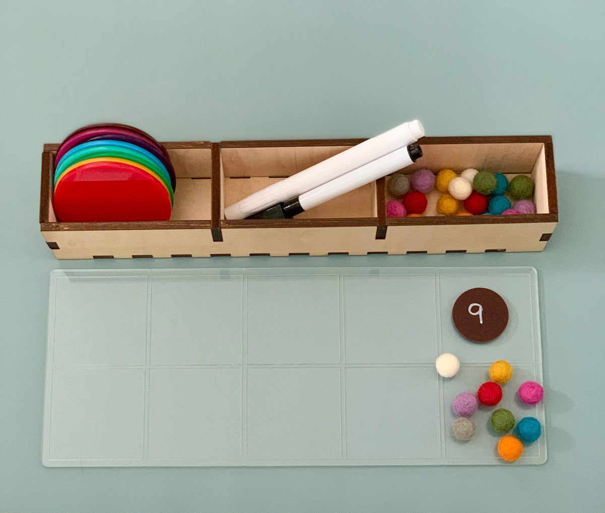 Wooden organizer holding colorful translucent discs, white markers, and multicolored felt balls on a light blue surface