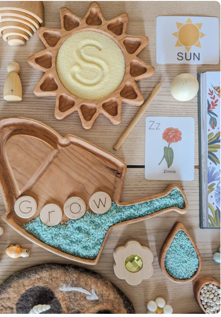 Wooden sun tray with yellow grains and watering can tray filled with blue rice spelling "Grow" on wooden table