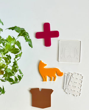 Glossy red cross, clear ribs plate, orange fox, clear xylophone with mallets, and brown bag shapes on white surface with green leafy plant nearby