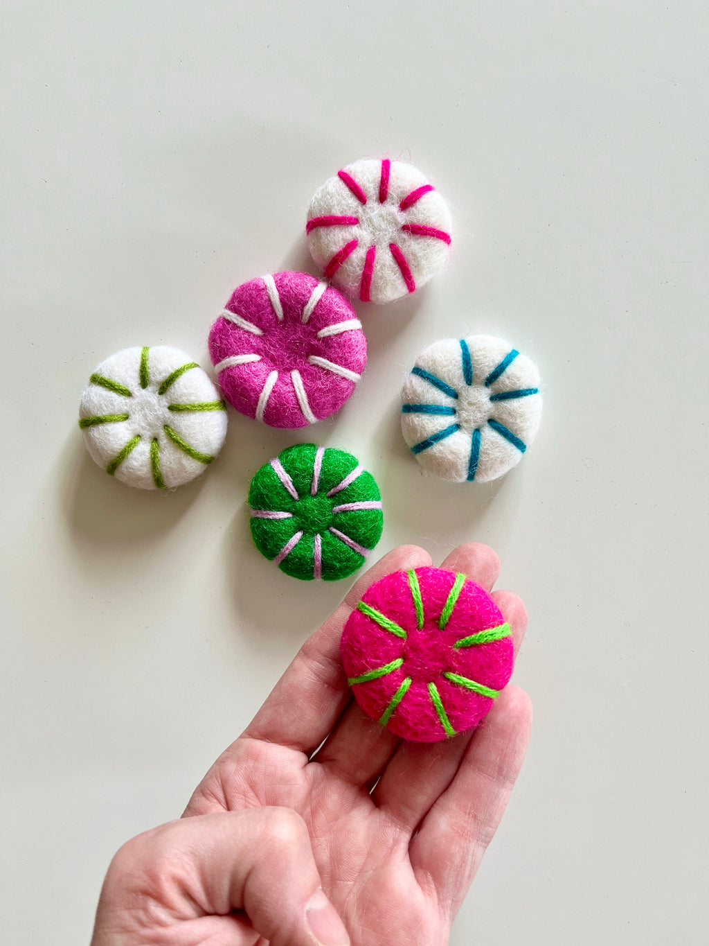 Hand holding bright pink felt circle with green stitched lines, surrounded by five similar felt circles on white surface